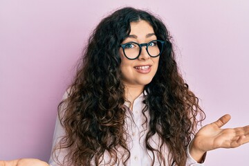 Young brunette woman with curly hair wearing casual clothes and glasses clueless and confused...