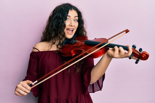 Young brunette musician woman with curly hair playing violin celebrating crazy and amazed for success with open eyes screaming excited.