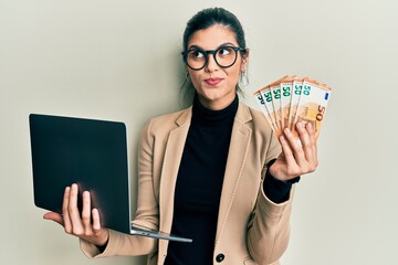Young hispanic woman wearing business style holding laptop and 50 euros smiling looking to the side...