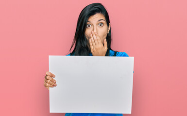 Young hispanic woman holding blank empty banner covering mouth with hand, shocked and afraid for mistake. surprised expression