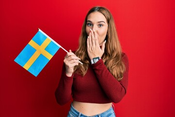 Young blonde woman holding sweden flag covering mouth with hand, shocked and afraid for mistake. surprised expression