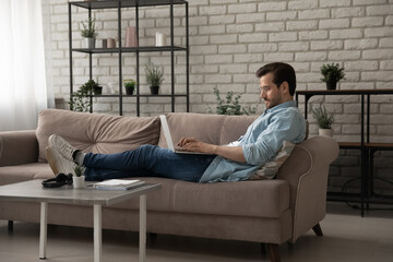 Side view confident man wearing glasses using laptop, lying on couch, spending leisure time, focused businessman or student looking at computer screen, freelancer working on online project