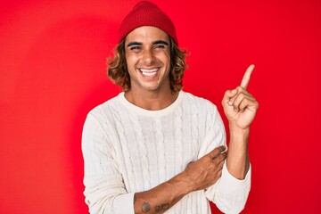 Young hispanic man wearing wool sweater and winter hat smiling happy pointing with hand and finger...