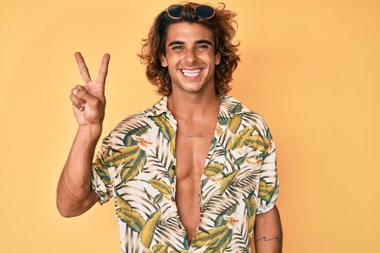Young hispanic man wearing summer shirt smiling with happy face winking at the camera doing victory sign. number two.