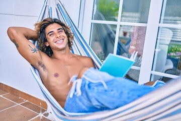 Young hispanic man relaxed reading book lying on the hammock at terrace.