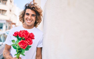 Young handsome hispanic man holding bouquet of roses leaning on the wall at street of city