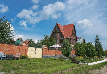 Fototapeta na wymiar A street in the village of Sedelnikovo (Ural, Russia) with a beautiful tall wooden house in the form of a tower, a fence and a green lawn with flower beds and a bench. Lifestyle, care and relaxation 