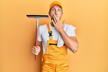 Young handsome man wearing glass clenaer uniform and squeegee covering mouth with hand, shocked and afraid for mistake. surprised expression