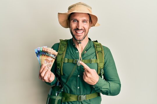Handsome man with beard wearing explorer hat holding canadian dollars smiling happy pointing with hand and finger