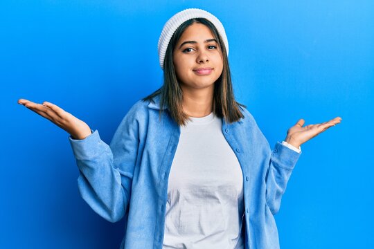 Young latin woman wearing cute wool cap clueless and confused expression with arms and hands raised. doubt concept.