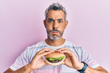 Middle age grey-haired man eating a tasty classic burger puffing cheeks with funny face. mouth inflated with air, catching air.