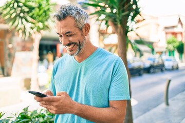 Middle age grey-haired man using smartphone at street of city looking positive and happy standing and smiling with a confident smile showing teeth
