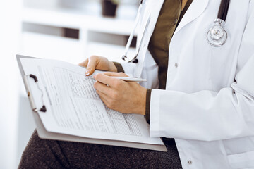 Unknown woman-doctor writing something at clipboard while sitting at the chair, close-up. Therapist wearing green blouse at work is filling up medication history record. Medicine concept