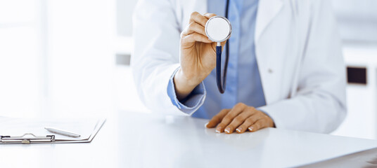 Unknown woman-doctor in blue blouse is holding stethoscope head while sitting at the desk in clinic, close-up. Physician ready to examine and help patient. Medicine concept