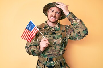 Young caucasian man wearing camouflage army uniform holding usa flag stressed and frustrated with hand on head, surprised and angry face
