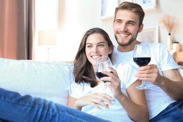 Happy couple sitting, relaxing on couch in living room, using laptop for online shopping together, watching movie, drinking red wine.