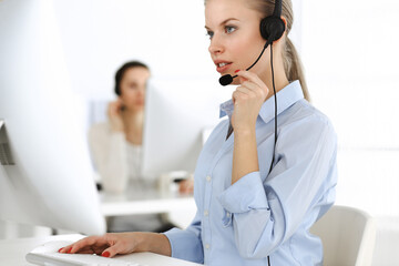 Blond woman call operator is using computer and headset for consulting clients online. Group of...