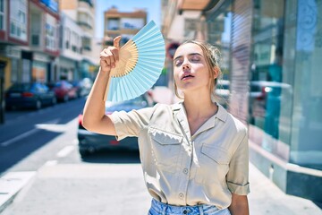Young beautiful blonde caucasian woman smiling happy outdoors on a sunny day using handfan for hot weather