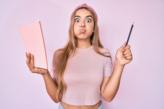 Young beautiful blonde woman holding book and pencil puffing cheeks with funny face. mouth inflated with air, catching air.