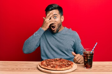 Handsome hispanic man eating tasty pepperoni pizza peeking in shock covering face and eyes with hand, looking through fingers afraid