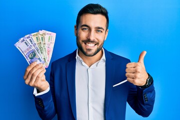 Young hispanic businessman wearing business suit holding indian rupee banknotes smiling happy and positive, thumb up doing excellent and approval sign