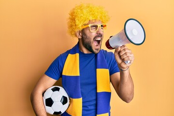 Young hispanic man football supporter holding soccer ball using megaphone angry and mad screaming frustrated and furious, shouting with anger looking up.