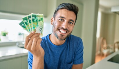 Young handsome man smiling happy holding israeli shekels banknotes at home