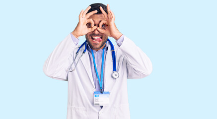 Young hispanic man wearing doctor uniform and stethoscope doing ok gesture like binoculars sticking tongue out, eyes looking through fingers. crazy expression.