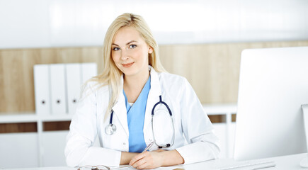 Woman-doctor at work while sitting at the desk in hospital or clinic. Blonde cheerful physician filling up medication history record form