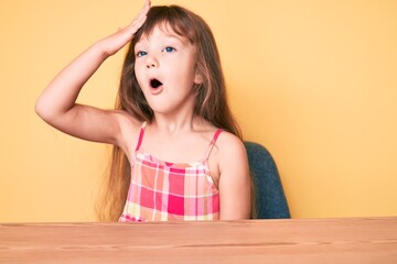 Little caucasian kid girl with long hair wearing casual clothes sitting on the table surprised with hand on head for mistake, remember error. forgot, bad memory concept.