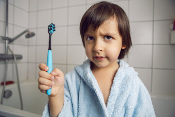 baby boy in a white bathroom stands in a blue robe and holds a toothbrush in his hand