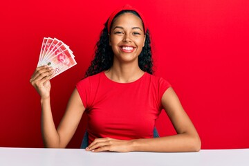 Young african american girl holding israel shekels sitting on the table looking positive and happy standing and smiling with a confident smile showing teeth