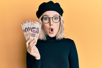 Young blonde girl holding mexican pesos scared and amazed with open mouth for surprise, disbelief face