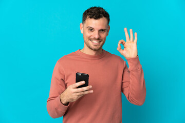 Young caucasian handsome man isolated on blue background using mobile phone and doing OK sign