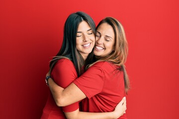 Hispanic family of mother and daughter wearing casual clothes over red background hugging oneself...