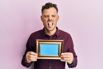 Young hispanic man holding empty frame sticking tongue out happy with funny expression.