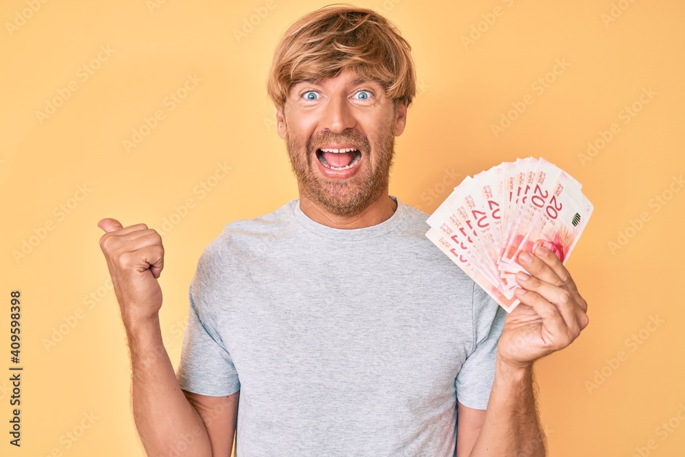 Poster Young blond man holding israeli shekels banknotes pointing thumb up to the side smiling happy with open mouth