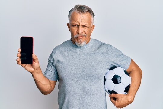 Middle age grey-haired man holding football ball showing smartphone skeptic and nervous, frowning upset because of problem. negative person.