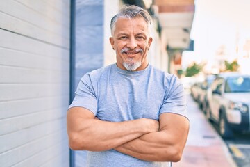 Middle age hispanic grey-haired man with crossed arms smiling happy at the city.