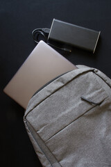 A gray textile city backpack with a silver laptop or ultrabook and an external battery on a black background. Traveling with gadgets.