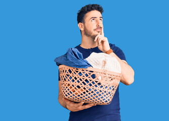 Young hispanic man holding laundry basket serious face thinking about question with hand on chin,...
