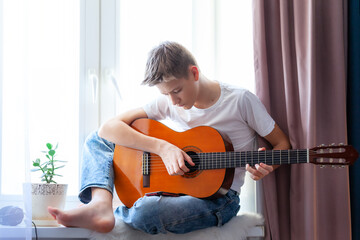 Handsome guy playing acoustic guitar while sitting on windowsill at home in his room