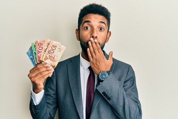 Handsome hispanic business man with beard holding canadian dollars covering mouth with hand, shocked and afraid for mistake. surprised expression