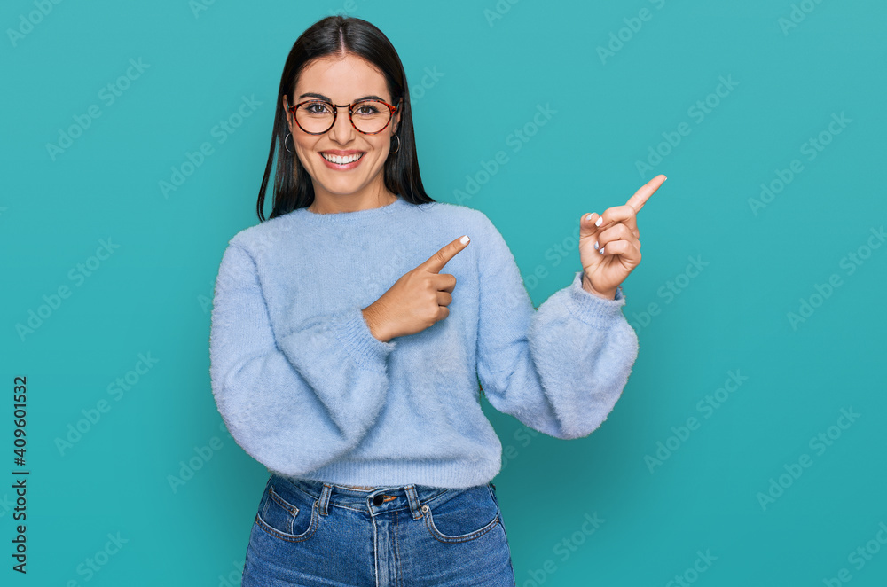 Wall mural Young hispanic woman wearing casual clothes and glasses smiling and looking at the camera pointing with two hands and fingers to the side.