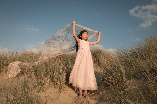 Fineart portrait of girl in long classic pink dress standing on dune holding white lace flying in the wind