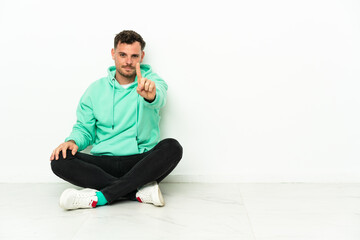 Young handsome caucasian man sitting on the floor showing and lifting a finger
