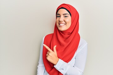 Young beautiful hispanic girl wearing traditional islamic hijab scarf cheerful with a smile of face pointing with hand and finger up to the side with happy and natural expression on face