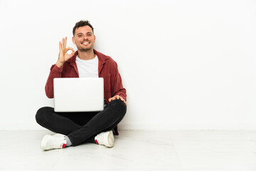 Young handsome caucasian man sit-in on the floor with laptop showing ok sign with fingers