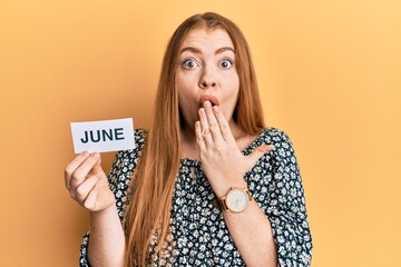 Young beautiful redhead woman holding june word on paper covering mouth with hand, shocked and...