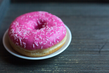 side view to bright pink donut on white round plate and aged wooden grey background. Isolated object close up. Traditional American sweets for breakfast. Copy space. White sugar decoration on top. 